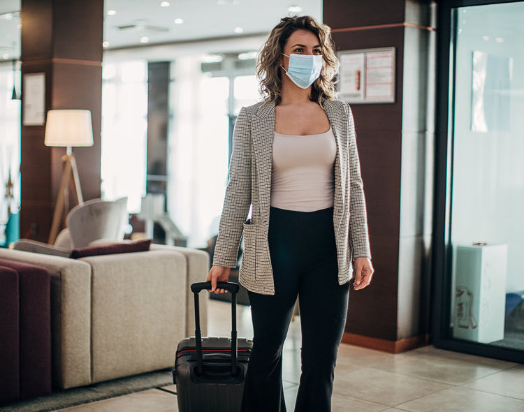 woman in face mask rolling luggage into hotel lobby