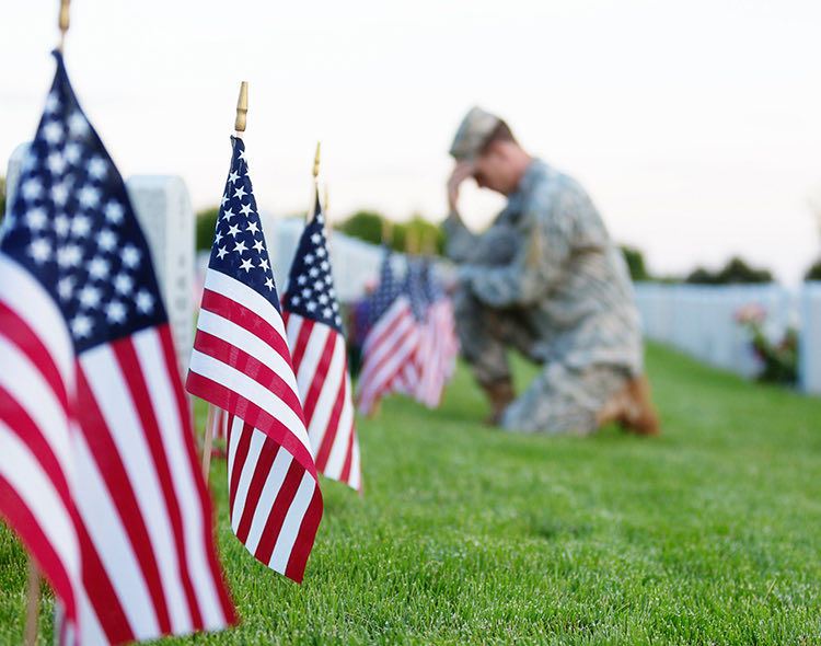 small american flags in ground at cemetery with a soldier kneeling at a gravesite