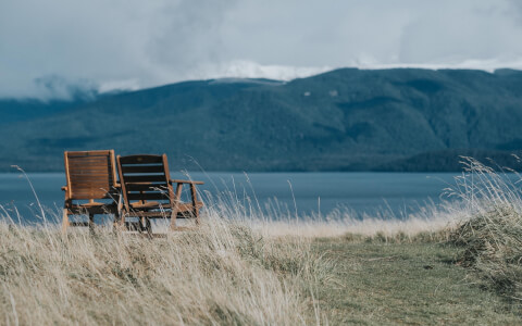 two wodden chairs at the shore of a lake with mountains in the background