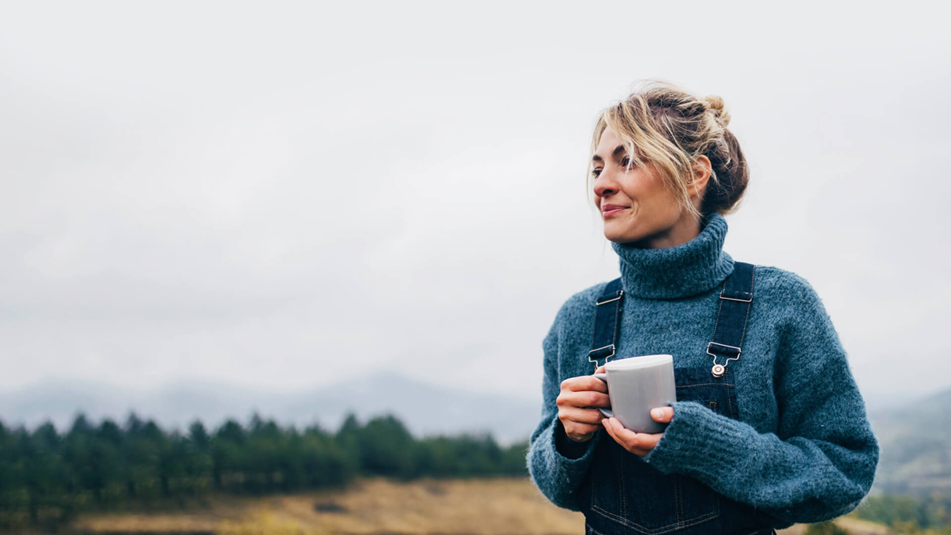 blonde woman holding a mug while standing outside in a sweater