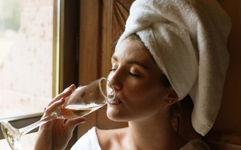 woman wearing a white shit with a towel on her head taking a sip of wine 
